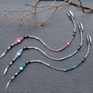 Può includere: Tre fermagli per capelli in macramè rosa, blu e verde acqua, ciascuno con un ciondolo a forma di piuma d'argento. I fermagli hanno un design intrecciato con perline nere e argentate e una clip di metallo nella parte superiore. Un rametto è sullo sfondo.