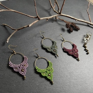 Puede incluir: Cuatro pares de pendientes de aro de macramé en colores lavanda, verde oliva, verde brillante y burdeos intenso. Cada pendiente presenta un aro de latón, intrincados patrones de cordón anudado y pequeñas cuentas metálicas.