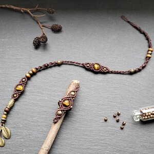 Peut inclure: Un accessoire de coiffure en macramé marron orné de pierres œil-de-tigre, de perles métalliques et d'une breloque feuille en laiton. Il comprend un pic à cheveux assorti avec une pierre centrale et un petit flacon en verre rempli de perles.