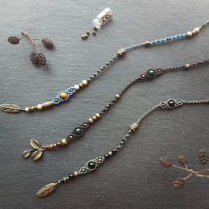 Könnte beinhalten: Drei Makramee-Armbänder mit geflochtenen Kordeln in Blau, Dunkelbraun und Olivgrün. Jedes Armband enthält einen zentralen Edelstein, kleine Metallperlen, einen kleinen Buddha-Kopf-Anhänger sowie einen hängenden Feder- oder Blattanhänger.
