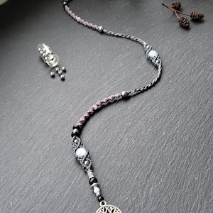 Peut inclure: Un collier macramé fait à la main avec un pendentif arbre de vie argenté. Le collier est composé d'un cordon noir, gris et lavande avec des perles blanches et noires. Une petite fiole en verre avec des perles argentées est en arrière-plan.