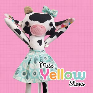 Puede incluir: Una muñeca de vaca de peluche hecha a mano con un patrón de manchas blancas y negras, que lleva una falda azul claro con un diseño floral y un lazo a juego. La muñeca tiene el texto "Miss Yellow Shoes" en la falda.
