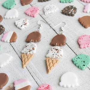 Puede incluir: Un par de pendientes colgantes con conos de helado de tres niveles de arcilla marr&oacute;n, blanca y marr&oacute;n moteada. Los conos est&aacute;n coronados con una bola de arcilla marr&oacute;n, blanca y rosa. Los pendientes est&aacute;n sobre una superficie de madera blanca con varios conos de helado peque&ntilde;os de arcilla en diferentes colores.