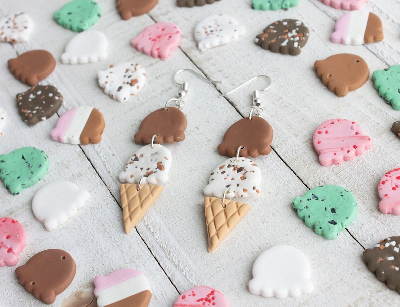 Puede incluir: Un par de pendientes colgantes con conos de helado de tres niveles de arcilla marr&oacute;n, blanca y marr&oacute;n moteada. Los conos est&aacute;n coronados con una bola de arcilla marr&oacute;n, blanca y rosa. Los pendientes est&aacute;n sobre una superficie de madera blanca con varios conos de helado peque&ntilde;os de arcilla en diferentes colores.
