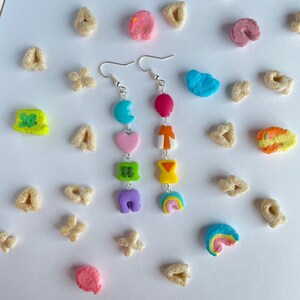 May include: A pair of dangle earrings with a silver hook and a chain with colorful charms. The charms are shaped like a crescent moon, heart, four-leaf clover, horseshoe, and rainbow. The charms are in shades of blue, pink, green, purple, and yellow.