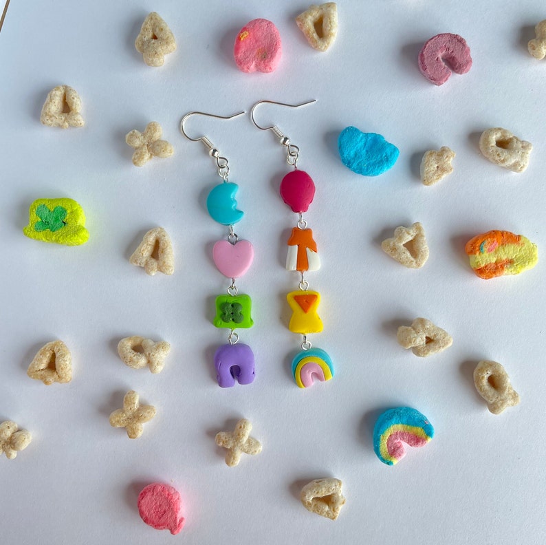 May include: A pair of dangle earrings with a silver hook and a chain with colorful charms. The charms are shaped like a crescent moon, heart, four-leaf clover, horseshoe, and rainbow. The charms are in shades of blue, pink, green, purple, and yellow.
