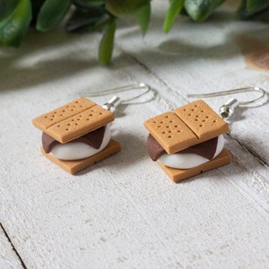 Puede incluir: Un par de pendientes colgantes con un mini s'more, hecho de arcilla polimérica marrón, blanca y beige. Los pendientes están hechos con ganchos de plata.
