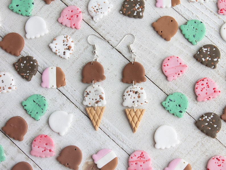 Puede incluir: Un par de pendientes colgantes con tres conos de helado apilados. Los conos est&aacute;n hechos de arcilla polim&eacute;rica y tienen un dise&ntilde;o marr&oacute;n, blanco con motas marrones y un remolino rosa. Los pendientes est&aacute;n sobre un fondo de madera blanco.