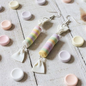 May include: A pair of candy-shaped earrings with pastel-colored stripes and white accents. The earrings are hanging from silver hooks and are surrounded by pastel-colored candies on a white wooden surface.