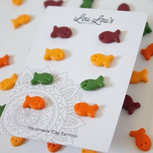 Op de afbeelding: Een set handgemaakte kleioorbellen in de vorm van goudviscrackers. De oorbellen zijn verkrijgbaar in vier kleuren: oranje, groen, rood en bruin. De oorbellen worden gepresenteerd op een witte kaart met de tekst "Lou Lou's Handmade Clay Earrings".