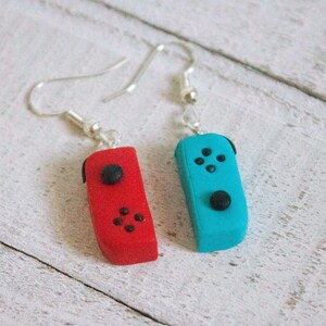 May include: A pair of handmade earrings featuring red and turquoise game controller designs. Each earring has a silver hook and a small black joystick and button details. The earrings are set against a white wooden background.