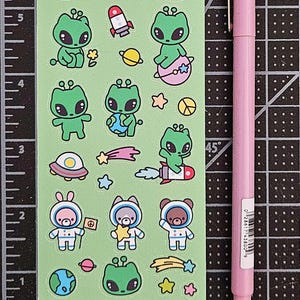 May include: A sheet of green stickers featuring cartoon aliens, space-themed elements, and animals in space suits. A pink pen is positioned next to the sticker sheet. The stickers are on a black cutting mat with inch measurements.