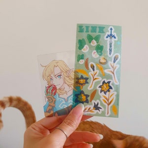 以下が含まれることがあります： 緑色の背景に、ゼルダの伝説シリーズのキャラクター、リンクの漫画イラストが描かれた透明なステッカーシート。ステッカーシートには、剣、花、その他の装飾的な要素が含まれています。シートの上部には緑色の文字で「LINK」と印刷されています。