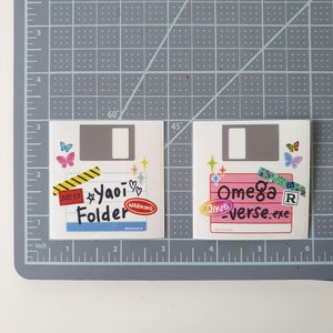 May include: Two white floppy disk stickers with colorful designs. The left sticker has a pink heart, stars, and the text "NC-17 Yaoi Folder" with a warning sign. The right sticker has a pink heart, stars, and the text "Omega love-verse.exe" with a red "R" rating.