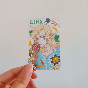 以下が含まれることがあります： ゼルダの伝説シリーズのビデオゲームから、リンクの漫画イラストが描かれたカード。リンクは金髪、青い目、緑色のチュニックを着ています。彼は赤いリンゴを手に持っています。カードは、黄色、緑、赤のアクセントがついた花柄のデザインです。カードの上部には黒い文字で「LINK」と印刷されています。