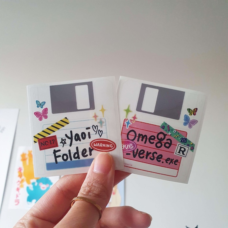 May include: Two colorful stickers with a floppy disk design. The first sticker is white with a black floppy disk and the text "Yaoi Folder" with a warning label. The second sticker is pink with a black floppy disk and the text "Omega -verse.exe".