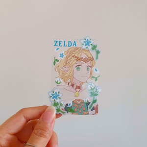 以下が含まれることがあります： ゼルダの伝説ビデオゲームシリーズのゼルダの漫画イラストが描かれた透明なステッカー。彼女はブロンドの髪と緑色の目をしており、緑と金の衣装を着ています。ステッカーは青い花と緑の葉で囲まれています。ステッカーの上部には青い文字で「ゼルダ」と印刷されています。