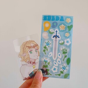 以下が含まれることがあります： 透明なプラスチックカードに、金髪緑目の女性キャラクターのイラストが描かれています。キャラクターは茶色と金の王冠と青と緑の衣装を着ています。カードの隣には、「ゼルダ」と印刷された青と白のステッカーシートがあります。ステッカーシートには、剣、花、葉が描かれています。