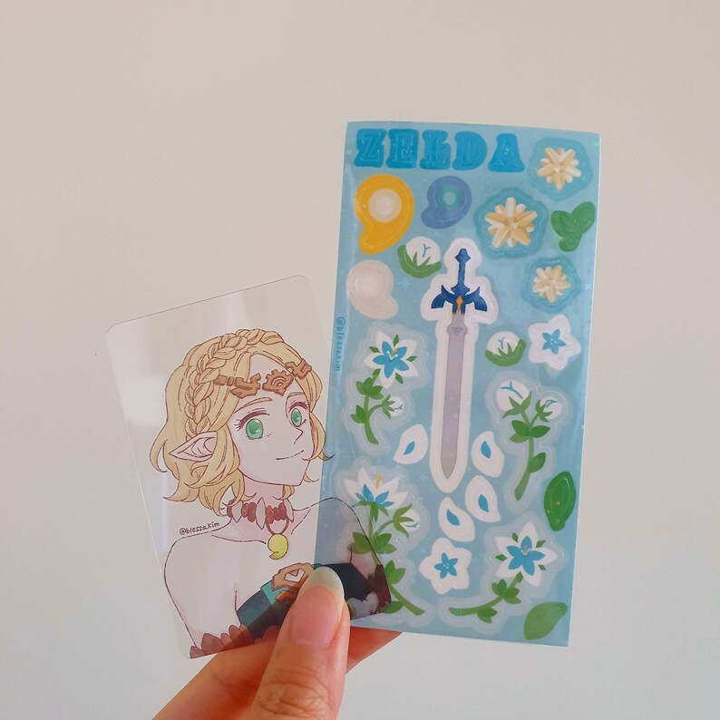 以下が含まれることがあります： 透明なプラスチックカードに、金髪緑目の女性キャラクターのイラストが描かれています。キャラクターは茶色と金の王冠と青と緑の衣装を着ています。カードの隣には、「ゼルダ」と印刷された青と白のステッカーシートがあります。ステッカーシートには、剣、花、葉が描かれています。