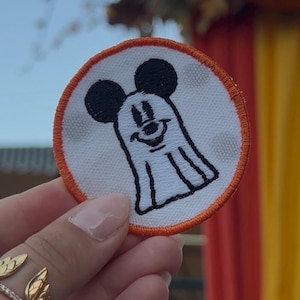 May include: A circular embroidered patch featuring a white ghost character with black outlines and black mouse ears. The patch has a bright orange stitched border and a white background with faint, light gray polka dots throughout the design.