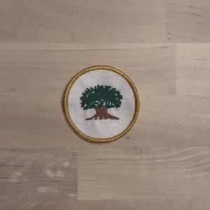May include: Circular embroidered patch featuring a green tree with a brown trunk and branches, set against a white background. The patch is bordered by a gold-colored trim. The design is centered on the patch.