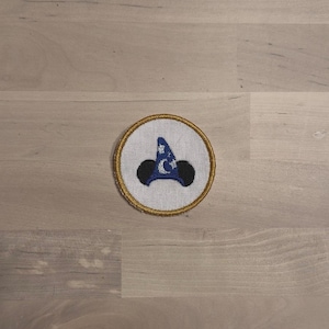 May include: Circular embroidered patch featuring a blue wizard hat with stars and a crescent moon, and black mouse ears. The design is on a white background with a gold border. The patch is likely for clothing or accessories.