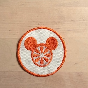 May include: Embroidered patch featuring a circular design with a white center and orange border. The center design is an orange Mickey Mouse head with a segmented orange design inside. The patch is likely for clothing or accessories.