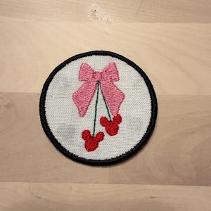 May include: Embroidered patch featuring a pink bow and two red cherries on a white background. The patch has a black border and is likely designed for clothing or accessories. The design is simple and cute.