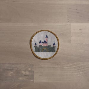 May include: Embroidered patch featuring a castle design. The castle has pink and gray walls, blue turrets, and a gold-colored border. The patch is circular and appears to be made of fabric.