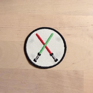 May include: Embroidered patch featuring a white circle with a black border. Inside, two crossed lightsabers are depicted, one red and one green. The lightsabers have silver handles. This patch is suitable for jackets, bags, or other fabric items.