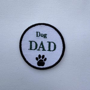 May include: A circular white patch with a black border. The patch features the words "Dog DAD" in green lettering, with a black paw print below the text. The patch is likely an embroidered item.