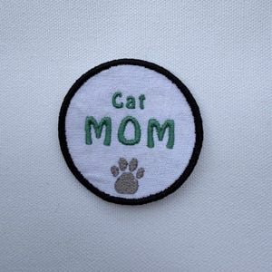 May include: A round, white patch with a black border. The patch features the words "Cat MOM" in green embroidered letters, with a gray paw print below. The patch is likely for clothing or accessories.