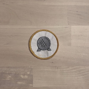 May include: Embroidered patch featuring a gray sphere with a crosshatch design, resting on two angled legs. The sphere is centered on a white circle, framed by a gold border. The patch is likely for clothing or accessories.