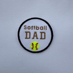 May include: Embroidered patch with the words "Softball DAD" in brown lettering, a yellow softball with red stitching, and a black border. The patch is circular and set against a white background.