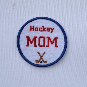 May include: Embroidered patch with a white background, a blue border, and the words "Hockey MOM" in red. Two crossed hockey sticks are at the bottom. The patch is circular.