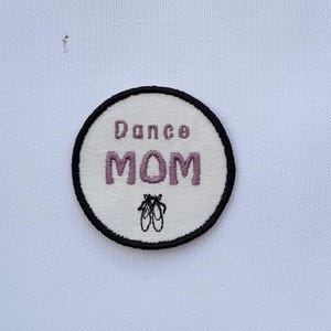 May include: A round embroidered patch with the words "Dance MOM" in purple thread. The patch has a white background with a black border and features a small illustration of ballet shoes.
