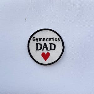 May include: A circular patch with the words "Gymnastics DAD" in black, embroidered on a white background. A red heart is below the word "DAD". The patch has a black border and is set against a white fabric.