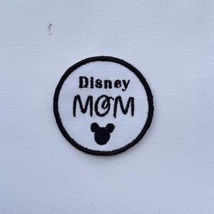 May include: A circular patch with the words "Disney MOM" embroidered in black on a white background. The patch has a black border and a small Mickey Mouse head icon at the bottom.