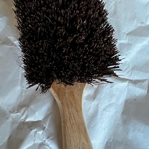 May include: A close-up of a cleaning brush with dark brown bristles and a light brown wooden handle. The handle has a curved design and a hole near the end. The bristles are densely packed.