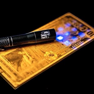 May include: A black flashlight with a silver patterned grip shines blue light on a gold-colored bill. The bill has the number 10 visible, and the flashlight has the letters "GB" on it.