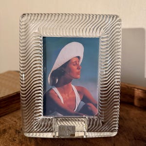 May include: Clear glass photo frame with a wavy design. The frame holds a color photograph of a person wearing a white hat and top. The brand name "MIKASA" is visible on the frame.