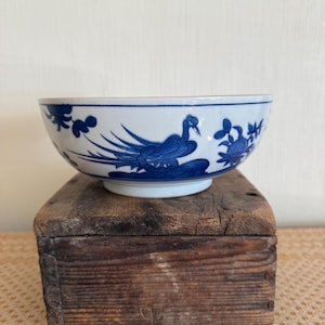 May include: A white and blue ceramic bowl with a bird and floral design. The bowl is set on a weathered wooden block. The bowl's rim and base are white, while the exterior features a detailed blue pattern.
