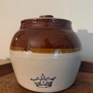 May include: A two-toned stoneware crock with a brown glazed upper section and a cream-colored lower body. The front features a blue stamped graphic of a crown with the number 2 inside. It includes a matching brown lid with a small knob handle.