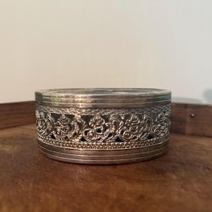 May include: A round silver-toned metal container featuring a decorative openwork filigree band around the center. The lid and base are finished with smooth, horizontal ridged borders, creating a structured, cylindrical appearance for this small box.