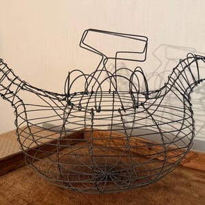 May include: A black wire hen-shaped basket with a handle. The basket is made of interwoven metal wires, forming the shape of a hen. It sits on a wooden surface, with a light-colored wall in the background. The basket is designed to hold eggs.