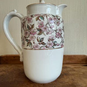 May include: White ceramic pitcher with a lid, handle, and spout. The upper portion is decorated with pink floral patterns and gold trim. The lower half is plain white. A decorative piece for serving beverages.