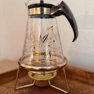 May include: A clear glass carafe featuring a gold botanical leaf pattern, a black handle, and a gold-toned metal band at the neck. The carafe rests on a gold-toned metal warming stand designed to hold a small candle or tea light beneath the base.