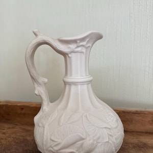 May include: A white ceramic pitcher with a curved handle and a spout. The pitcher features a detailed floral relief design on the body and handle. The pitcher is sitting on a wooden surface.