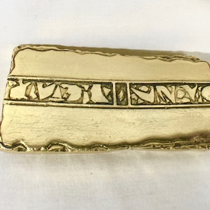 Könnte beinhalten: Ein goldfarbener Metallstab mit einem kunstvollen Design, das in die Oberfläche geätzt ist. Das Design ist ein sich wiederholendes Muster aus stilisierten Formen und Linien.