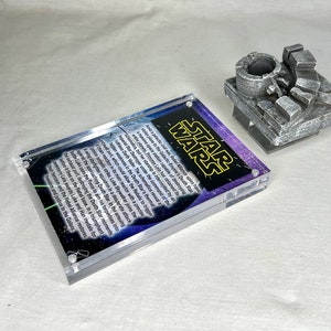 Peut inclure: Un cadre en acrylique transparent avec un fond noir et les mots "STAR WARS" en jaune. Le cadre contient une image imprimée d'une scène spatiale avec une citation sur l'Étoile de la Mort.